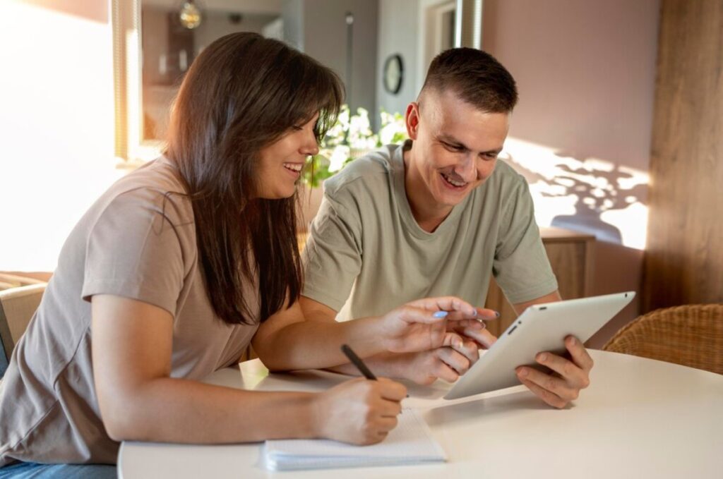 Two people smiling and interacting with a tablet at a bright, sunlit table.