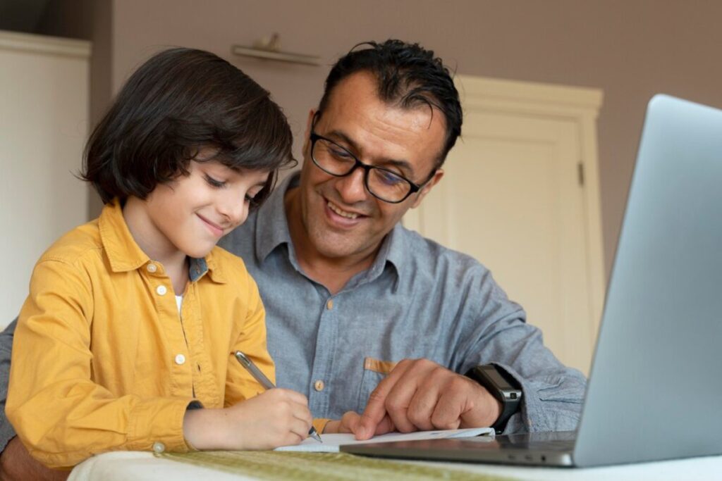 A man and a young boy happily engaged in writing and studying together in front of a laptop.