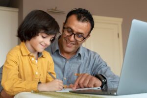 A man and a young boy happily engaged in writing and studying together in front of a laptop.