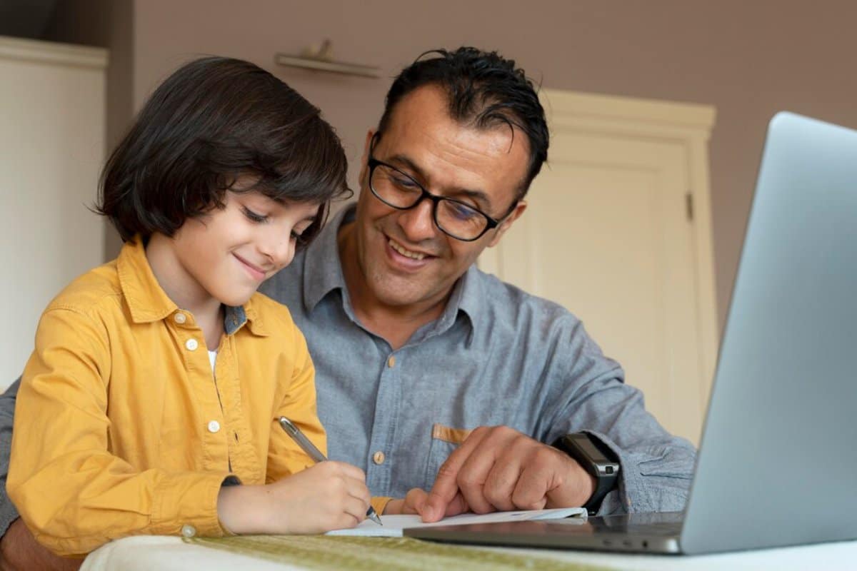 A man and a young boy happily engaged in writing and studying together in front of a laptop.