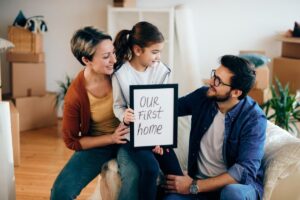 Family with a young girl holding a sign that reads "OUR FIRST HOME" in a living room filled with moving boxes.