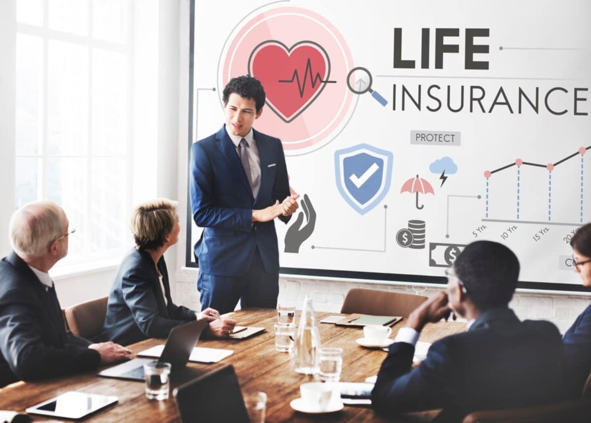 A man in a business suit presents life insurance concepts to colleagues in a meeting room with graphical display.