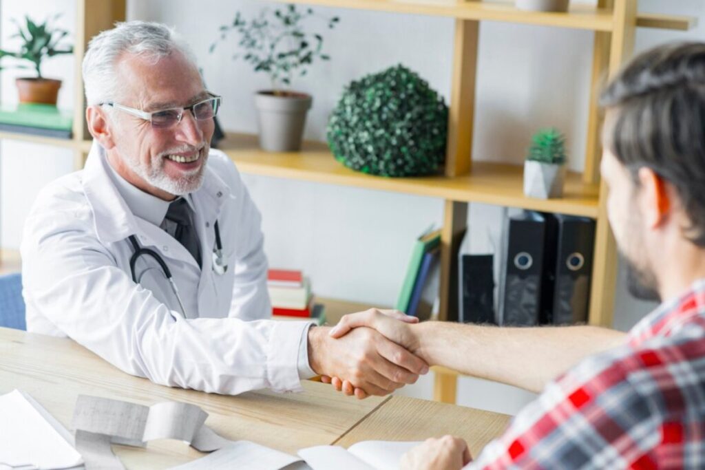 Senior male doctor shaking hands with a younger male patient at a desk in a bright office.
