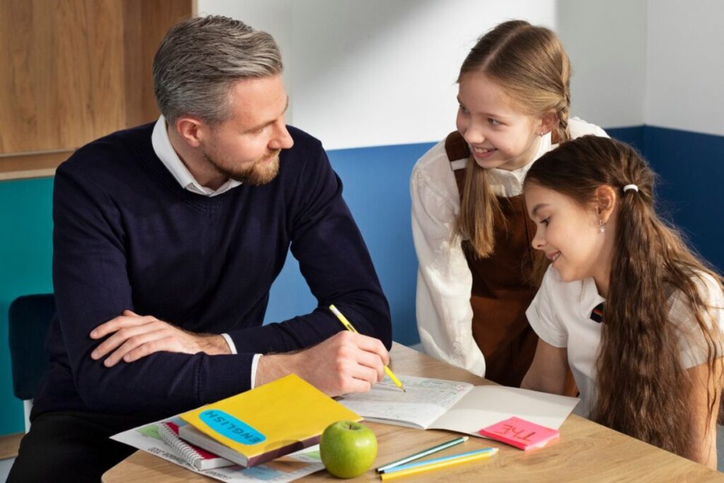 A man helps two young girls with schoolwork at a table with books and an apple.