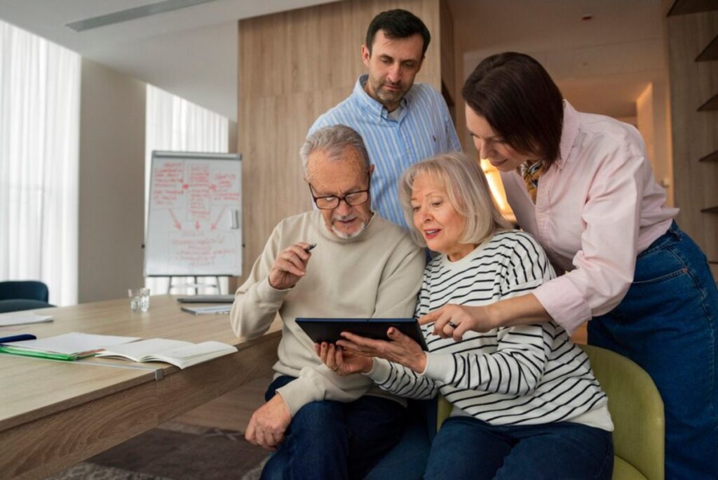 Two senior adults and two middle-aged adults look at a tablet together in a modern office setting.