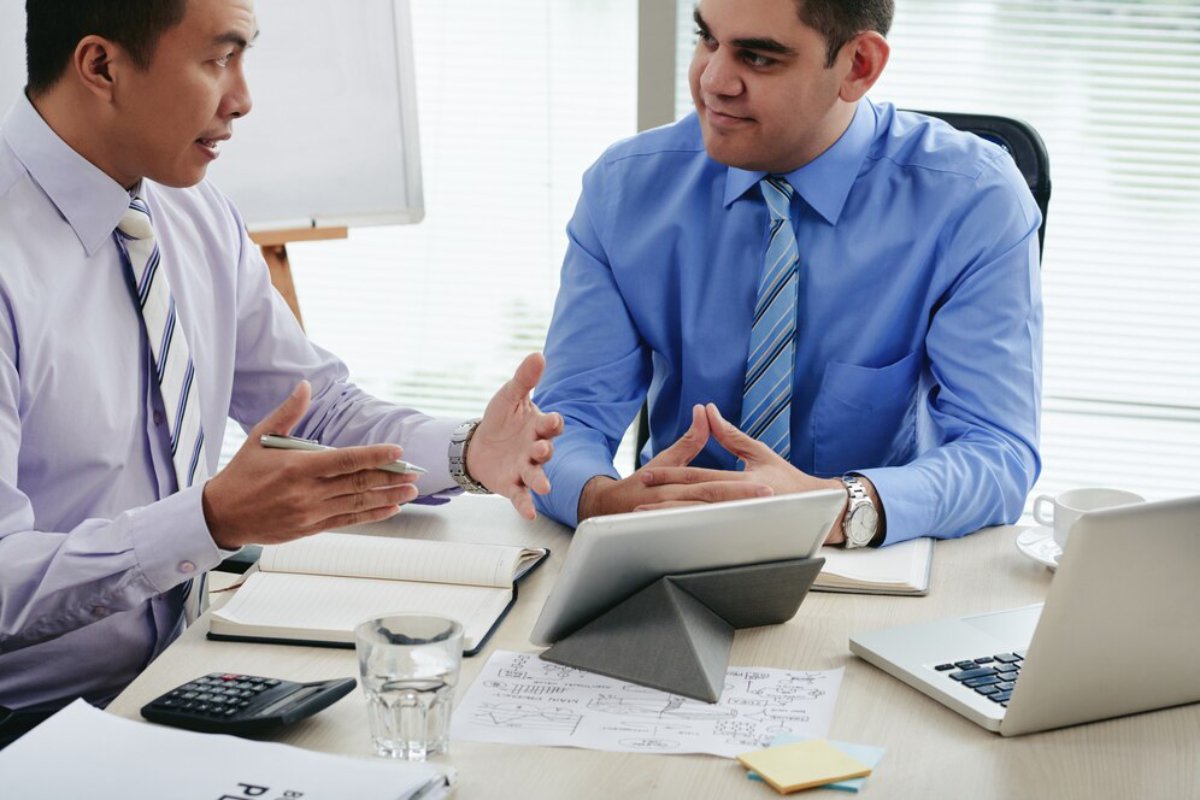 Two men in business attire engaged in a discussion at a desk with laptops, papers, and digital devices.