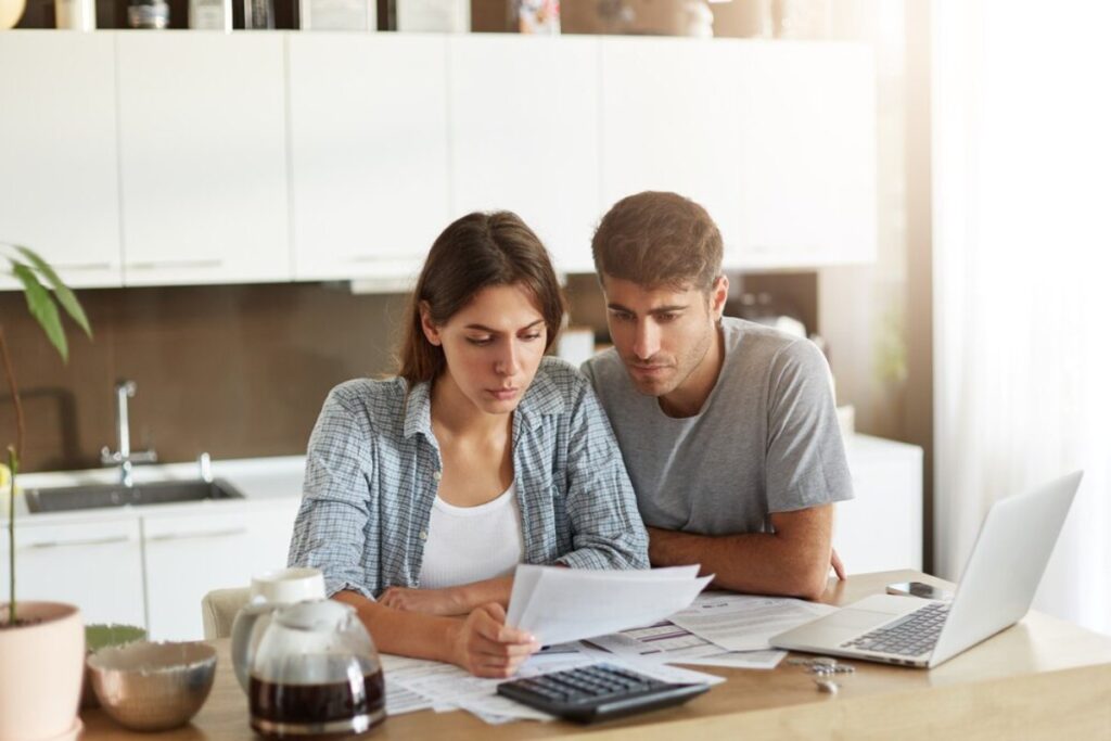 A couple reviews financial documents together in a bright kitchen with a laptop and calculator nearby.