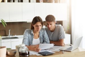 A couple reviews financial documents together in a bright kitchen with a laptop and calculator nearby.