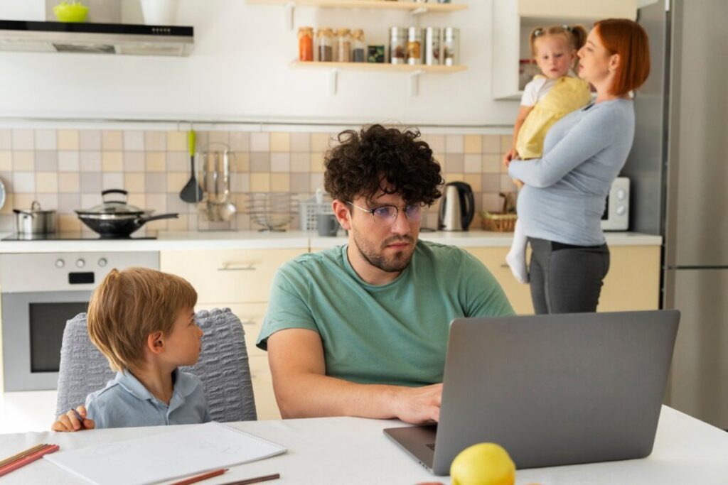 Man works on laptop in kitchen while talking to his son, as a woman holds a toddler in the background.