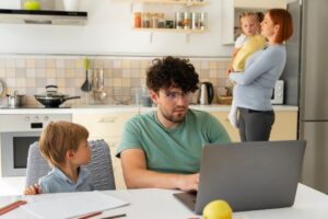 Man works on laptop in kitchen while talking to his son, as a woman holds a toddler in the background.