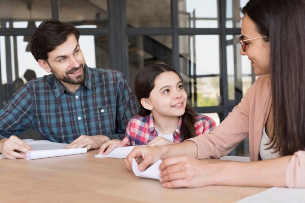 A family sits at a table, with a young girl smiling at her mother while the father watches affectionately.
