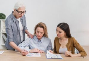 Three women of varying ages examine documents on a table, engaged in a discussion in an office setting.