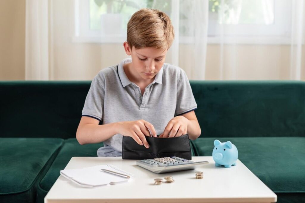 Young boy counting coins from wallet at table with calculator, notebook, and piggy bank nearby.