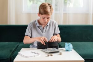 Young boy counting coins from wallet at table with calculator, notebook, and piggy bank nearby.