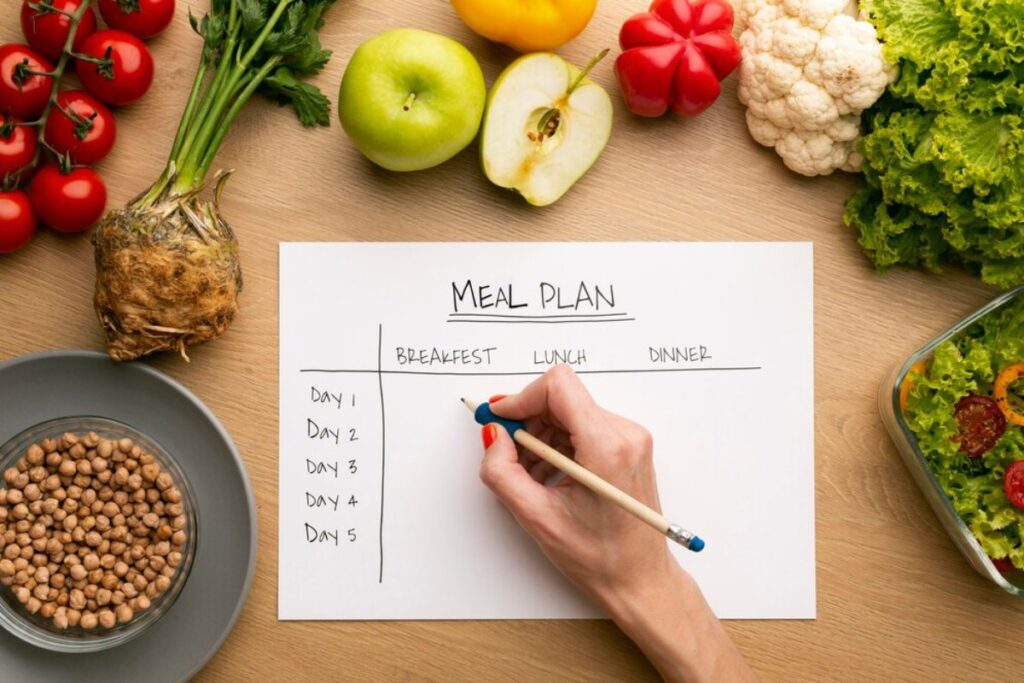A person's hand writing a meal plan with fresh vegetables and fruits around on a wooden table.