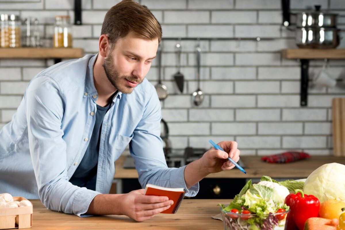 Man writing in a notebook and looking at a recipe in a modern kitchen with fresh vegetables on the table.