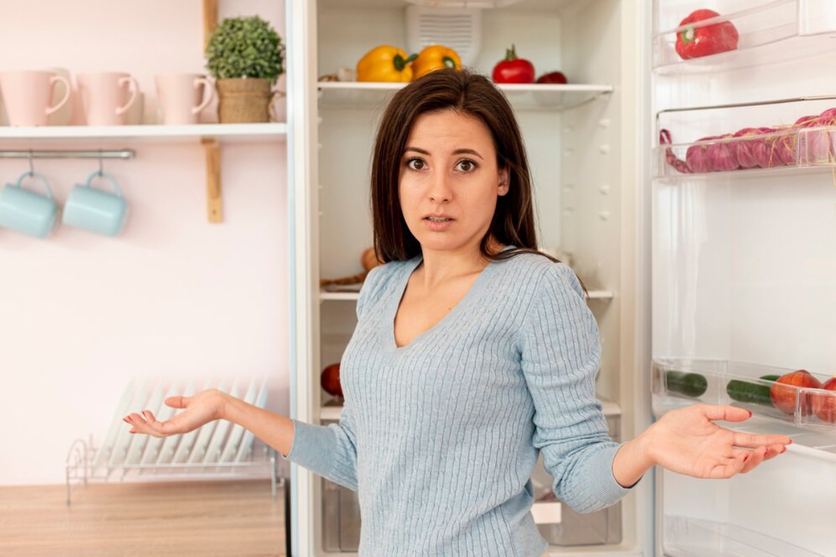 Surprised woman in a blue sweater, gesturing with open hands, standing by an open fridge stocked with vegetables.