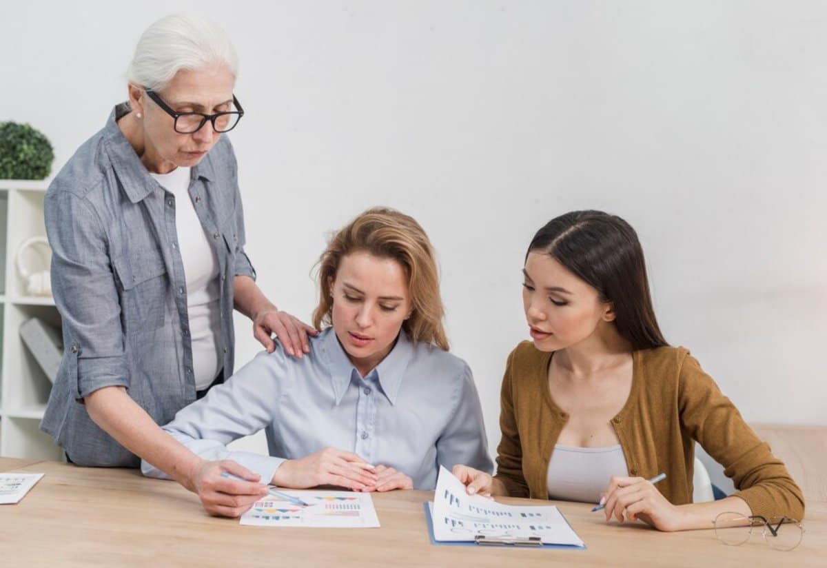 Three women of varying ages examine documents on a table, engaged in a discussion in an office setting.