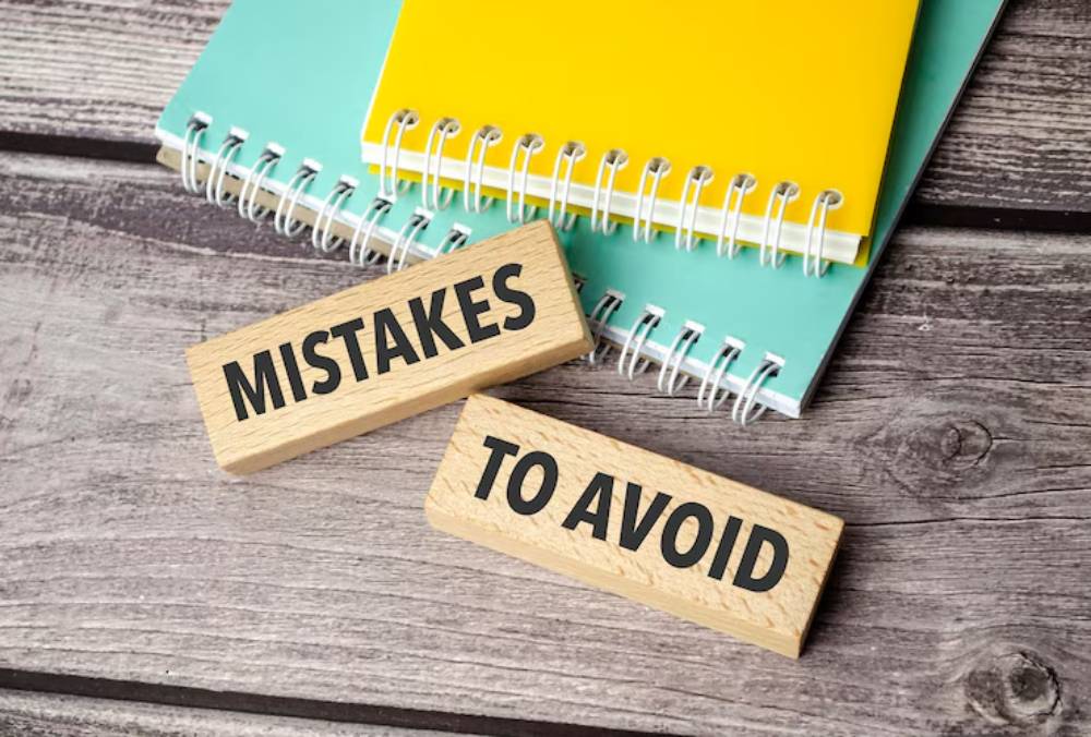 Two wooden blocks labeled MISTAKES and TO AVOID resting on a wooden surface, with colorful spiral notebooks in the background.