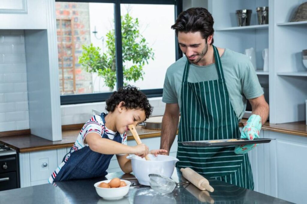 A child and an adult cook together in a bright kitchen, using a rolling pin and mixing ingredients in a bowl. Eggs are on the counter.