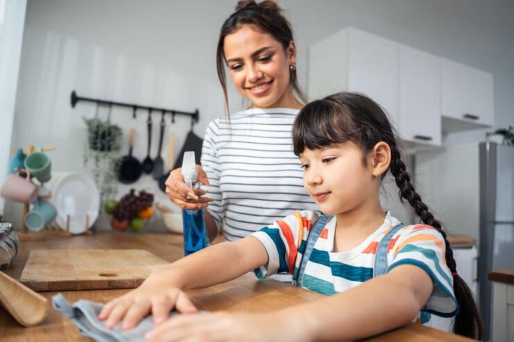 A woman and a girl are in a kitchen, with the girl reaching out towards a cloth while the woman holds a spray bottle.