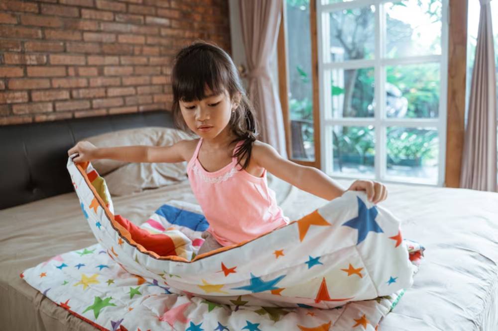 A child in a pink top is playfully unfolding a colorful star-patterned bedding on a neatly made bed in a cozy, bright room.