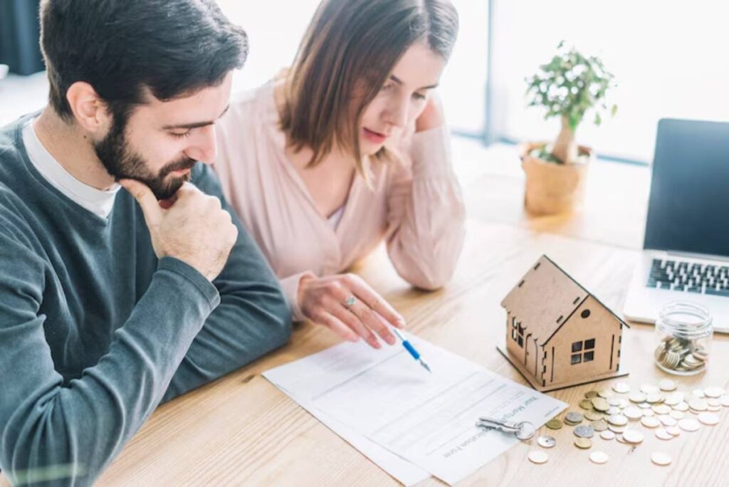 A couple discusses paperwork related to buying a home, with a small wooden house model and coins on the table beside a laptop.
