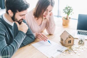 A couple discusses paperwork related to buying a home, with a small wooden house model and coins on the table beside a laptop.