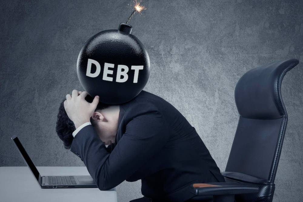 A stressed man in a suit sits at a desk, head down, with a bomb labeled DEBT about to explode above him.