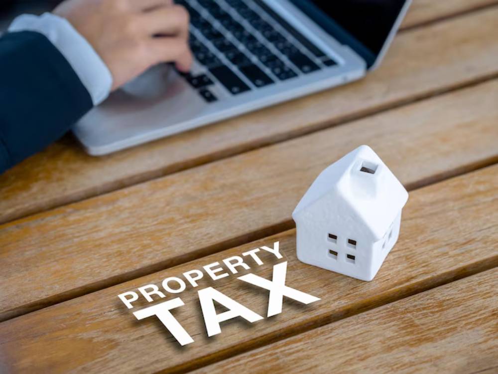A person using a laptop, with a white house model and the text PROPERTY TAX on a wooden table, representing tax-related activities.
