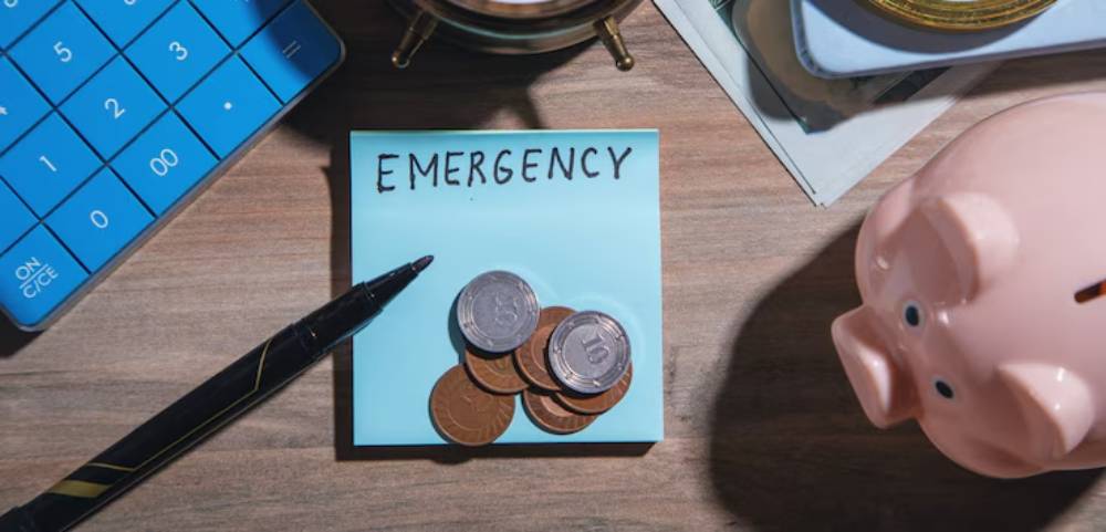 A blue sticky note labeled EMERGENCY with coins sits on a wooden desk beside a black pen, calculator, and a pink piggy bank.