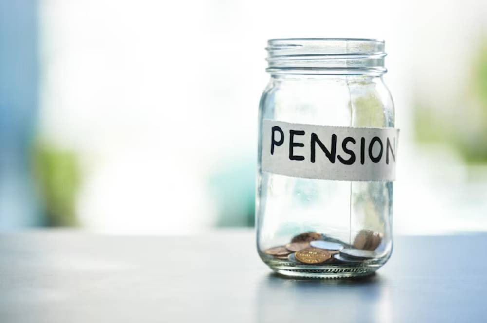 A glass jar labeled PENSION sits on a table, partly filled with coins, symbolizing savings for retirement.