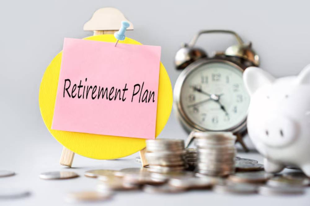 A sticky note labeled Retirement Plan on an easel, surrounded by coins, a piggy bank, and an alarm clock.