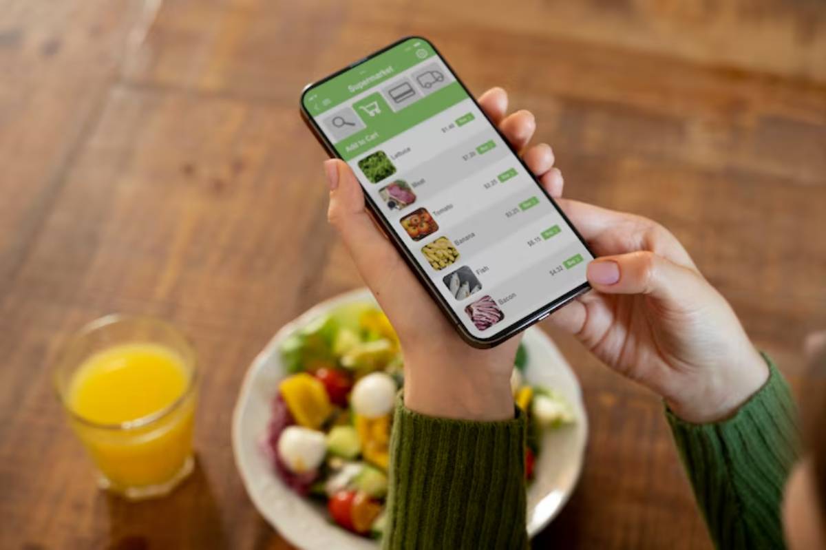 A person holds a smartphone displaying a grocery app, with a bowl of fresh salad and a glass of orange juice nearby.