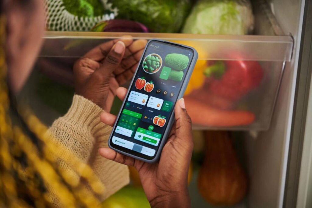 A person checks a smartphone app displaying grocery items while reaching into a fridge filled with fresh vegetables.