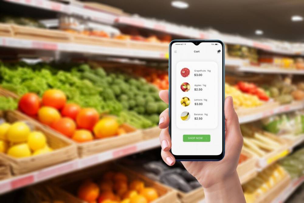 A hand holding a smartphone displays a shopping cart with fruit prices, amidst a colorful grocery produce aisle.