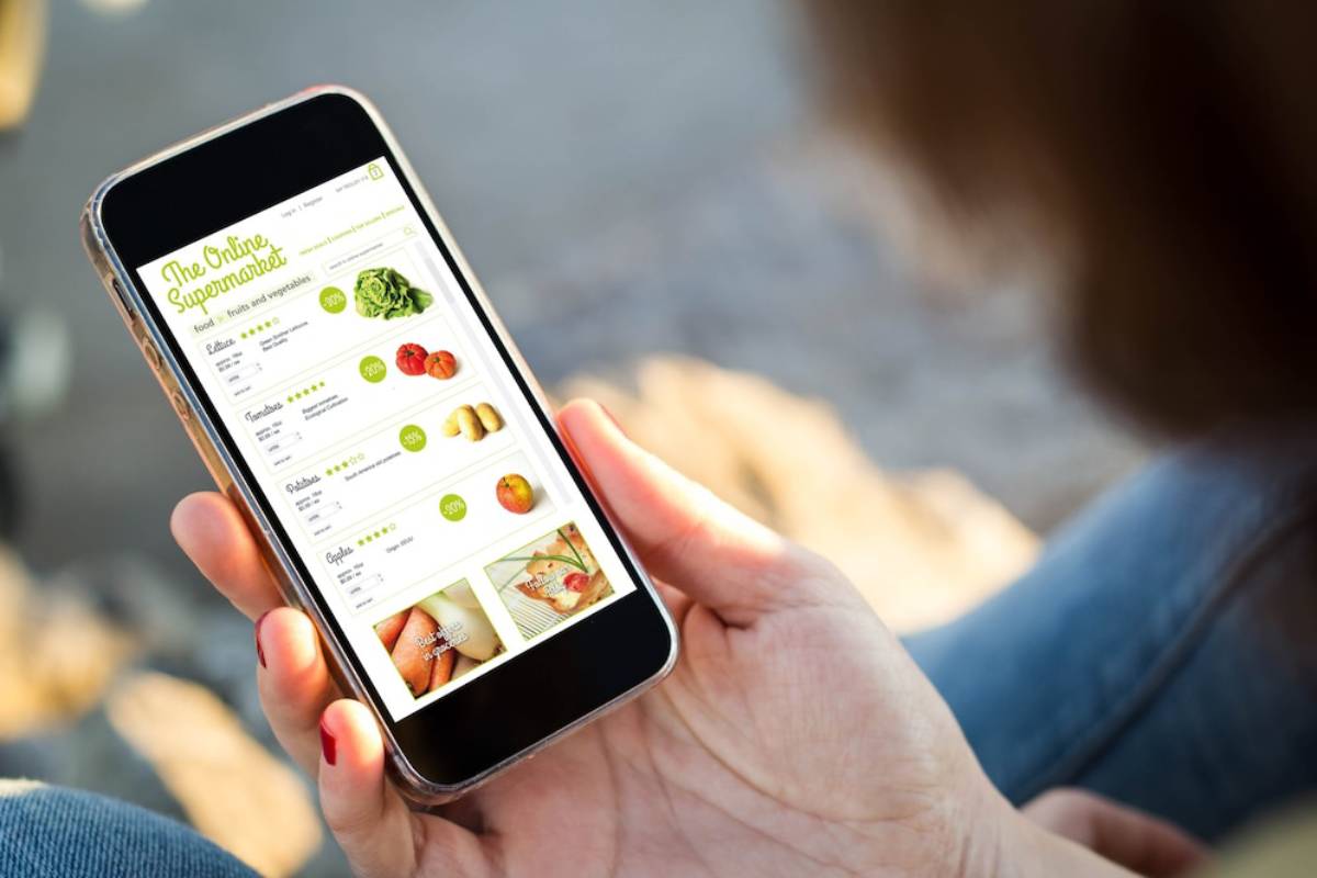 A person holds a smartphone displaying an online supermarket interface with various fruits and vegetables available for purchase.