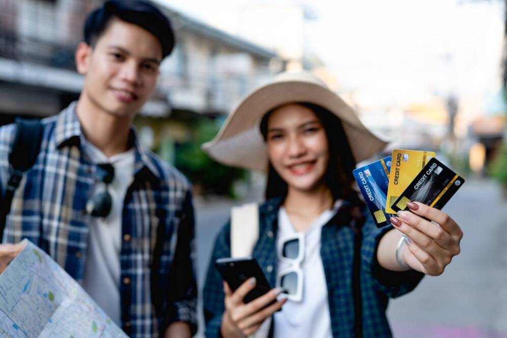 A young couple stands in a street, holding a map and multiple credit cards.