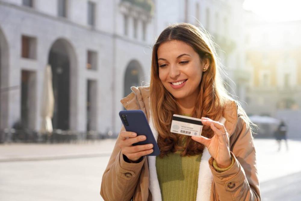 A woman in a casual coat holds a phone in one hand and a credit card in the other, standing on a sunlit street.